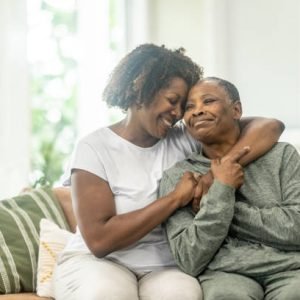 A middle aged woman of African decent wraps her arm around her Mother as she embraces her and they pose for a portrait.  They are both dressed comfortably and are laughing as they enjoy their time together.